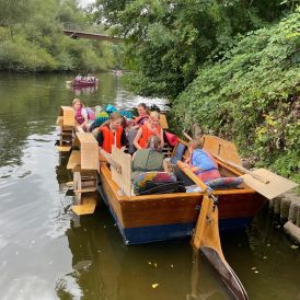Ein selbstgebautes Tretboot schwimmt auf einem Fluss nahe des Ufers. Mehrere Mädchen* und Frauen* sitzen mit Schwimmwesten im Boot. Ein Mädchen* lenkt das Boot.