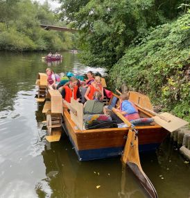 Ein selbstgebautes Tretboot schwimmt auf einem Fluss nahe des Ufers. Mehrere Mädchen* und Frauen* sitzen mit Schwimmwesten im Boot. Ein Mädchen* lenkt das Boot.